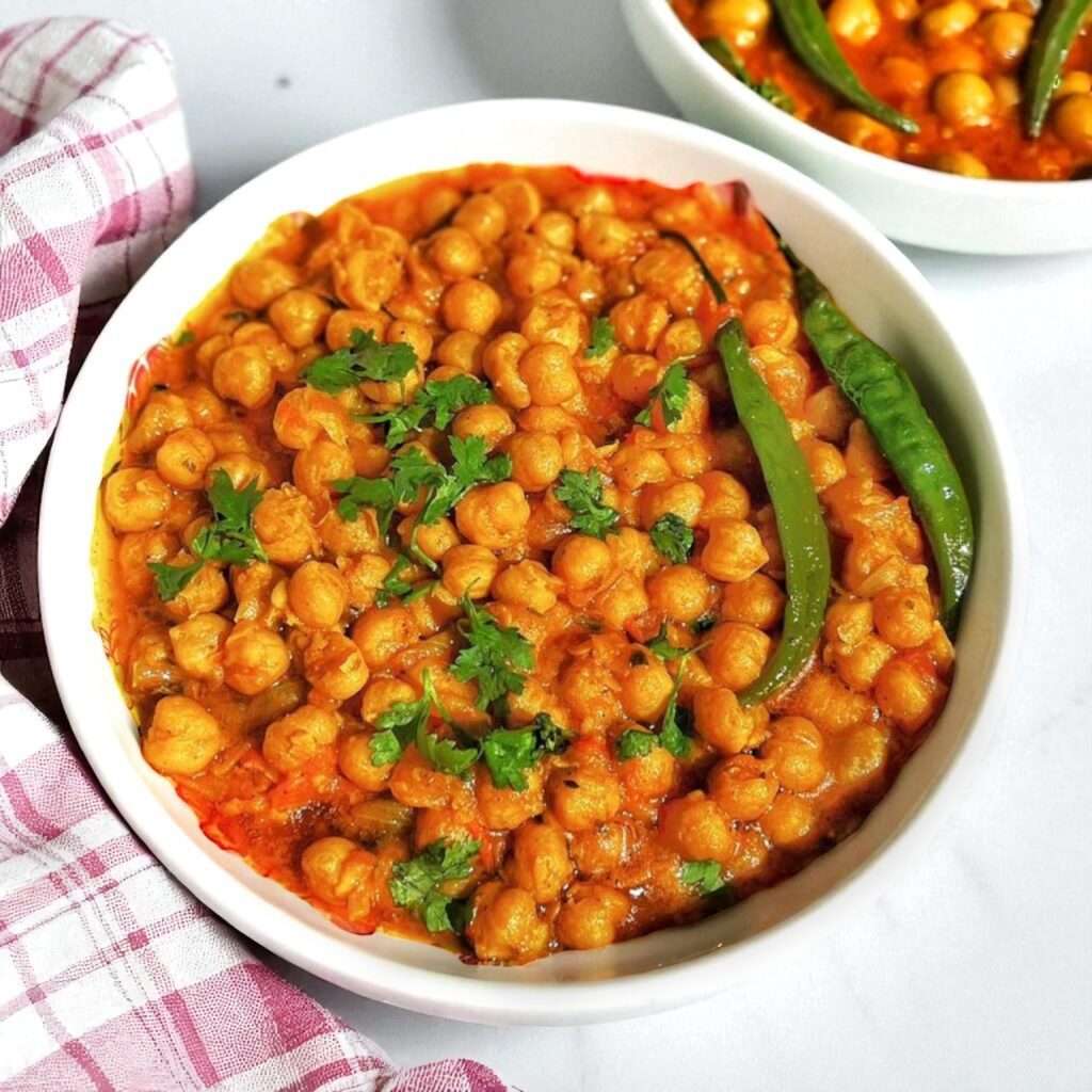 A bowl of chana masala (chickpea curry) arranged in ceramic bowl, garnished with cilantro and green chilies, served on a white marble surface.