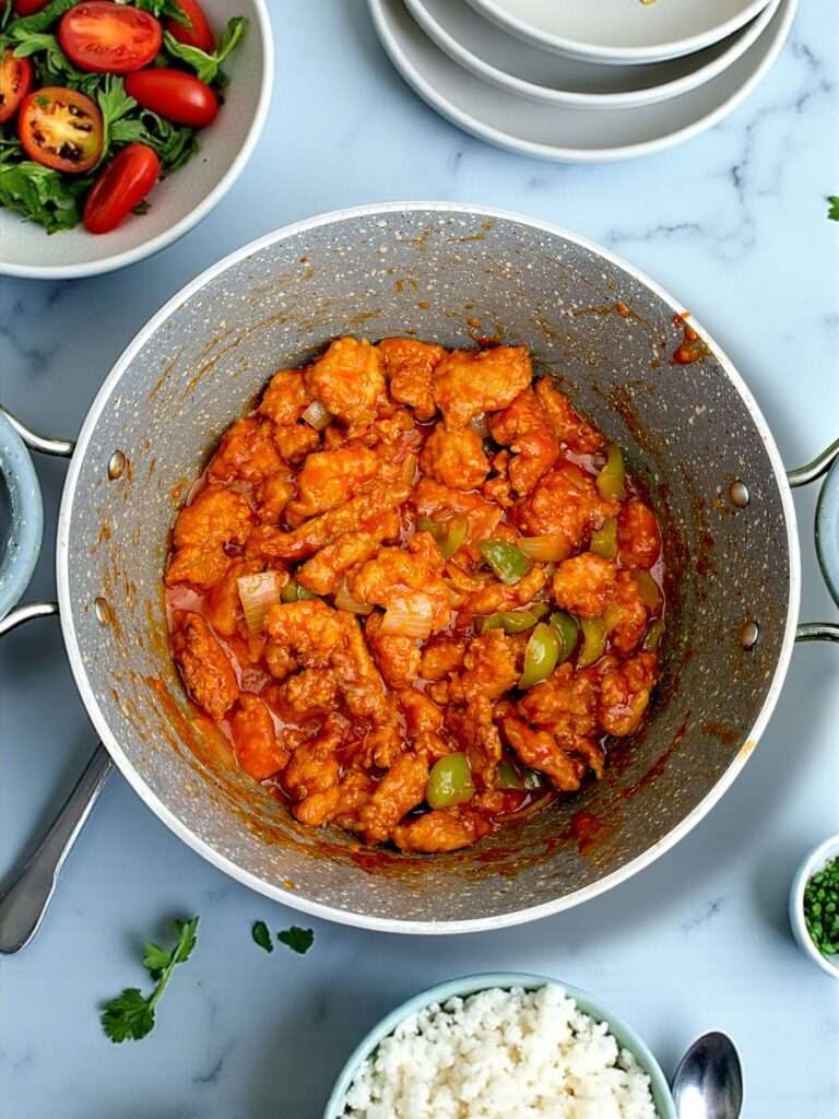Chicken Manchurian arranged in red handled marble pot, placed on white marble countertop, creating fine dining atmosphere.