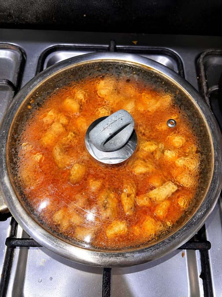 Chicken pieces simmering in a rich tomato curry under a covered pan
